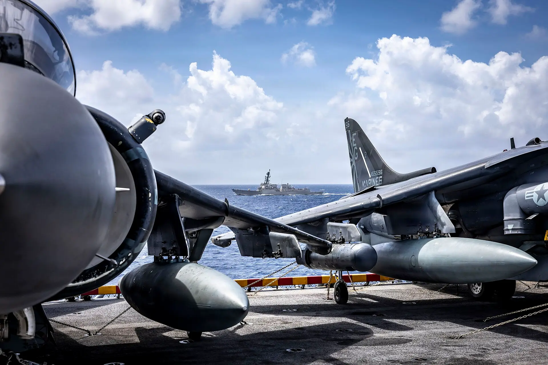 A U.S. Navy destroyer sails between two Harrier jets parked on the deck of an amphibious assault ship.