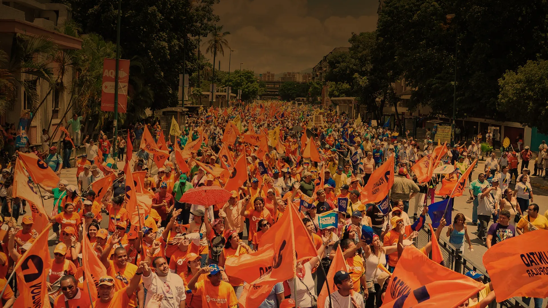 A crowd of Voluntad Popular supporters with orange flags fills a street.