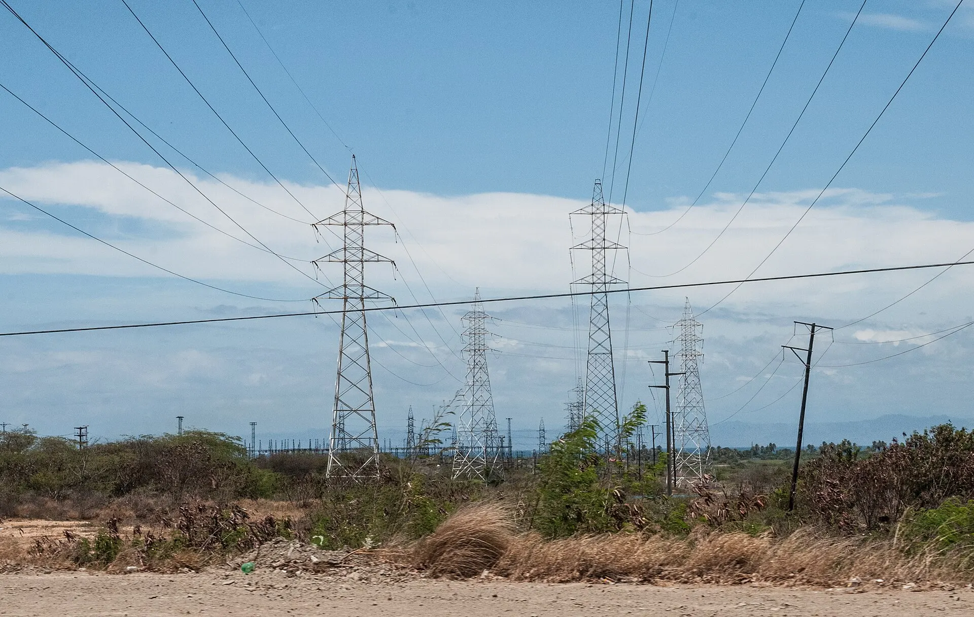 Steel electricity transmission pylons and power lines cross dry scrubland under a cloudy sky in Venezuela.