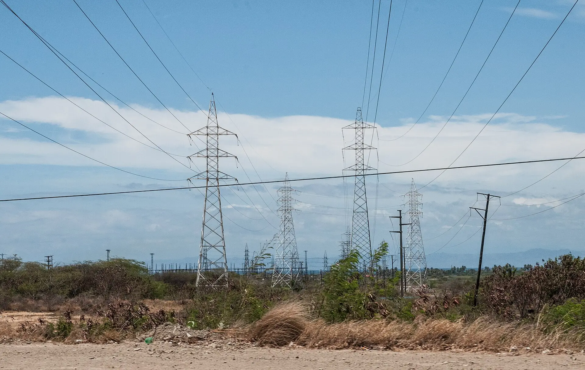Steel electricity transmission pylons and power lines cross dry scrubland under a cloudy sky in Venezuela.