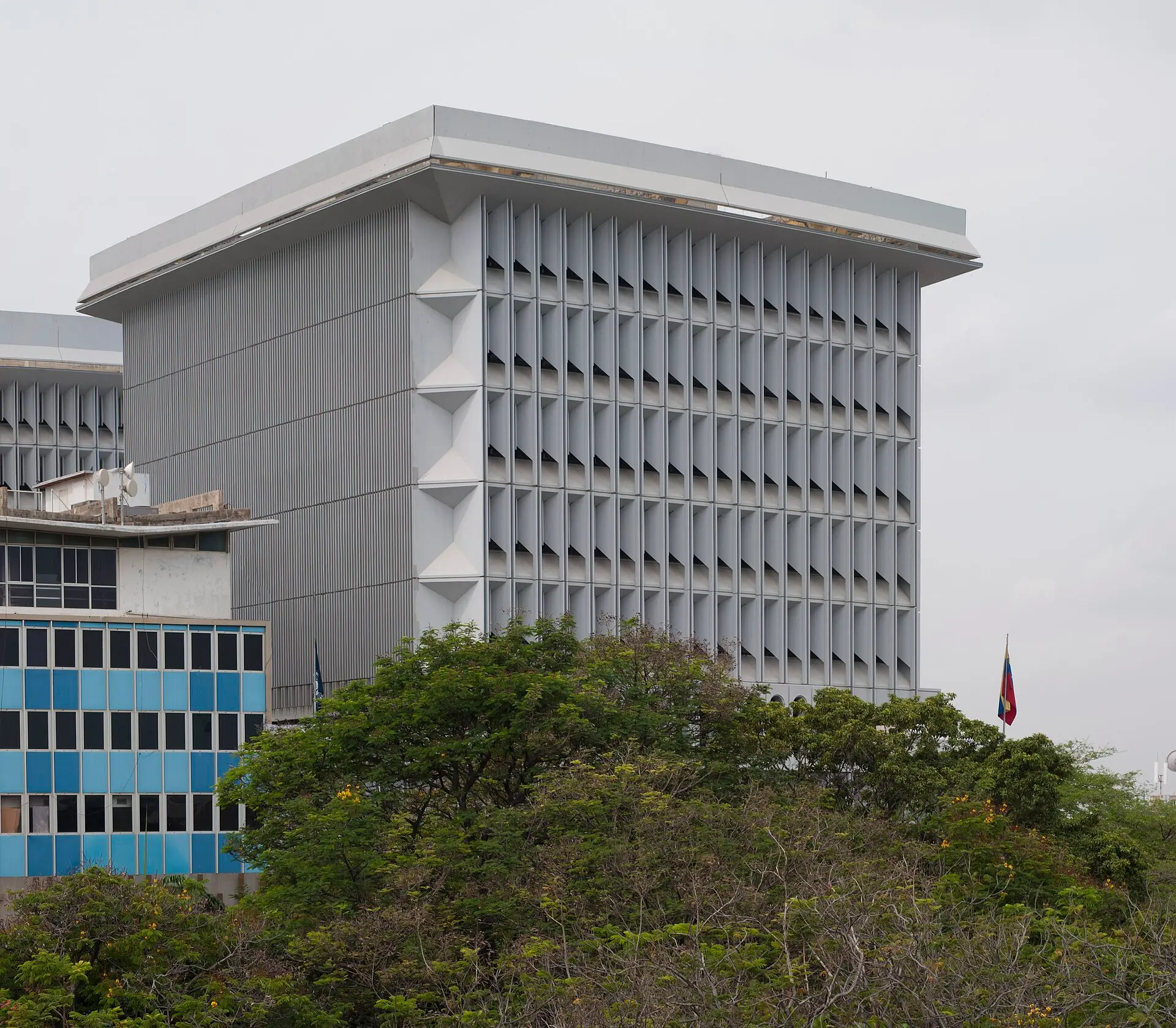 The grey modernist tower of the Central Bank of Venezuela in Maracaibo rises above trees, with a Venezuelan flag at right.