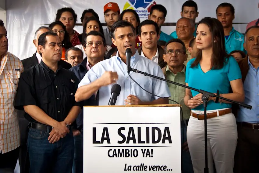 Leopoldo López speaks at a podium marked "La Salida, cambio ya!", with María Corina Machado and other politicians behind him.