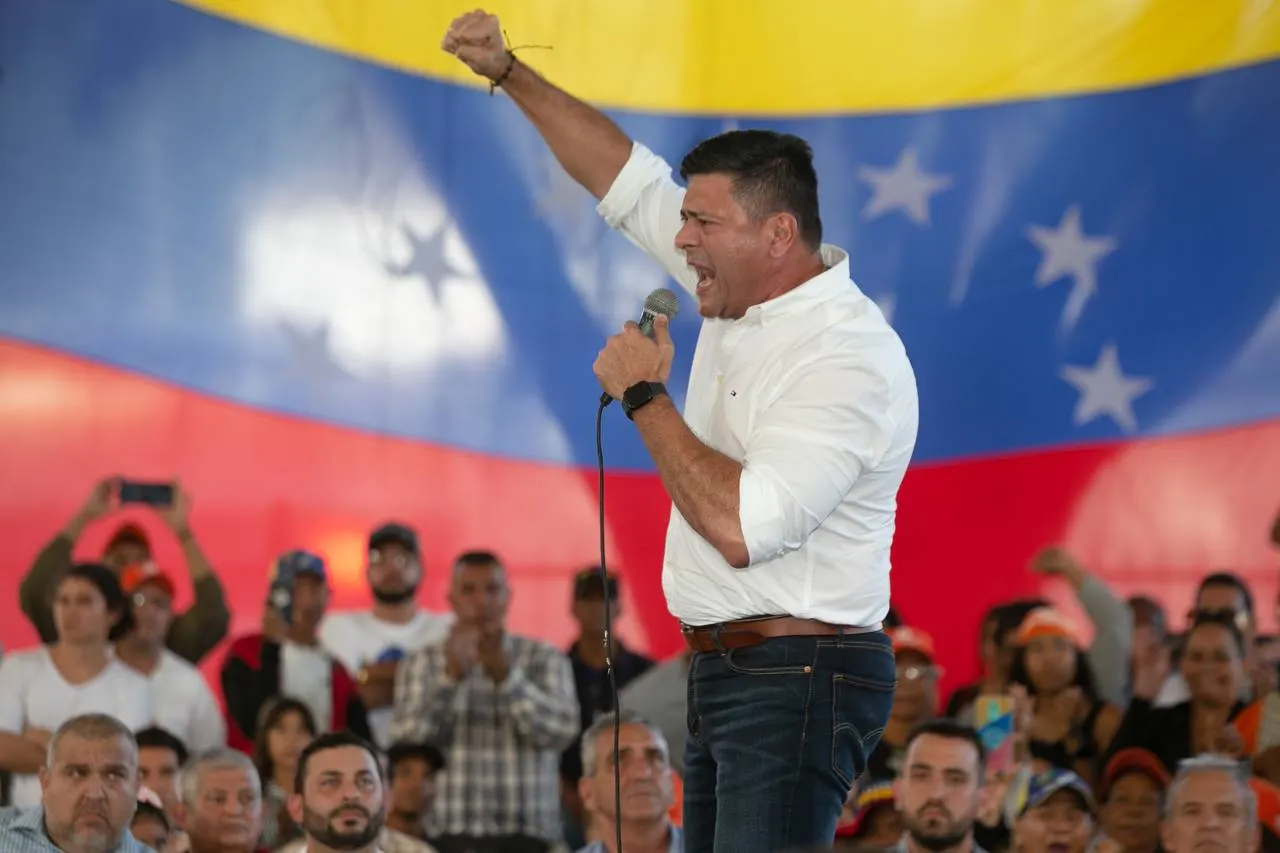 A speaker raises his fist before a crowd, in front of a large Venezuelan flag.