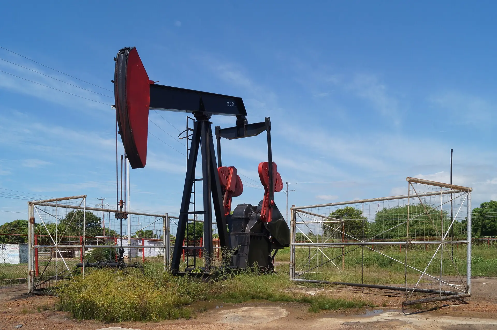 A red and blue oil pumpjack inside a wire fence in an oil field in Zulia, Venezuela, under a blue sky.