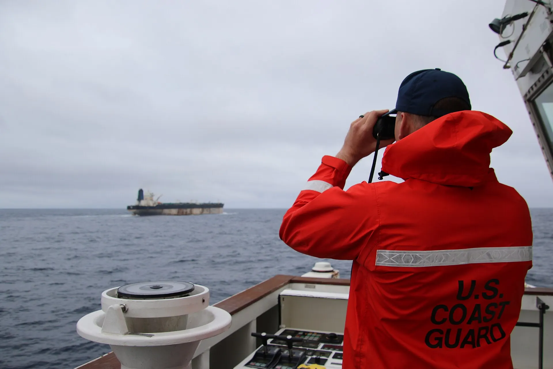 A U.S. Coast Guard crew member watches an oil tanker through binoculars from a cutter at sea.