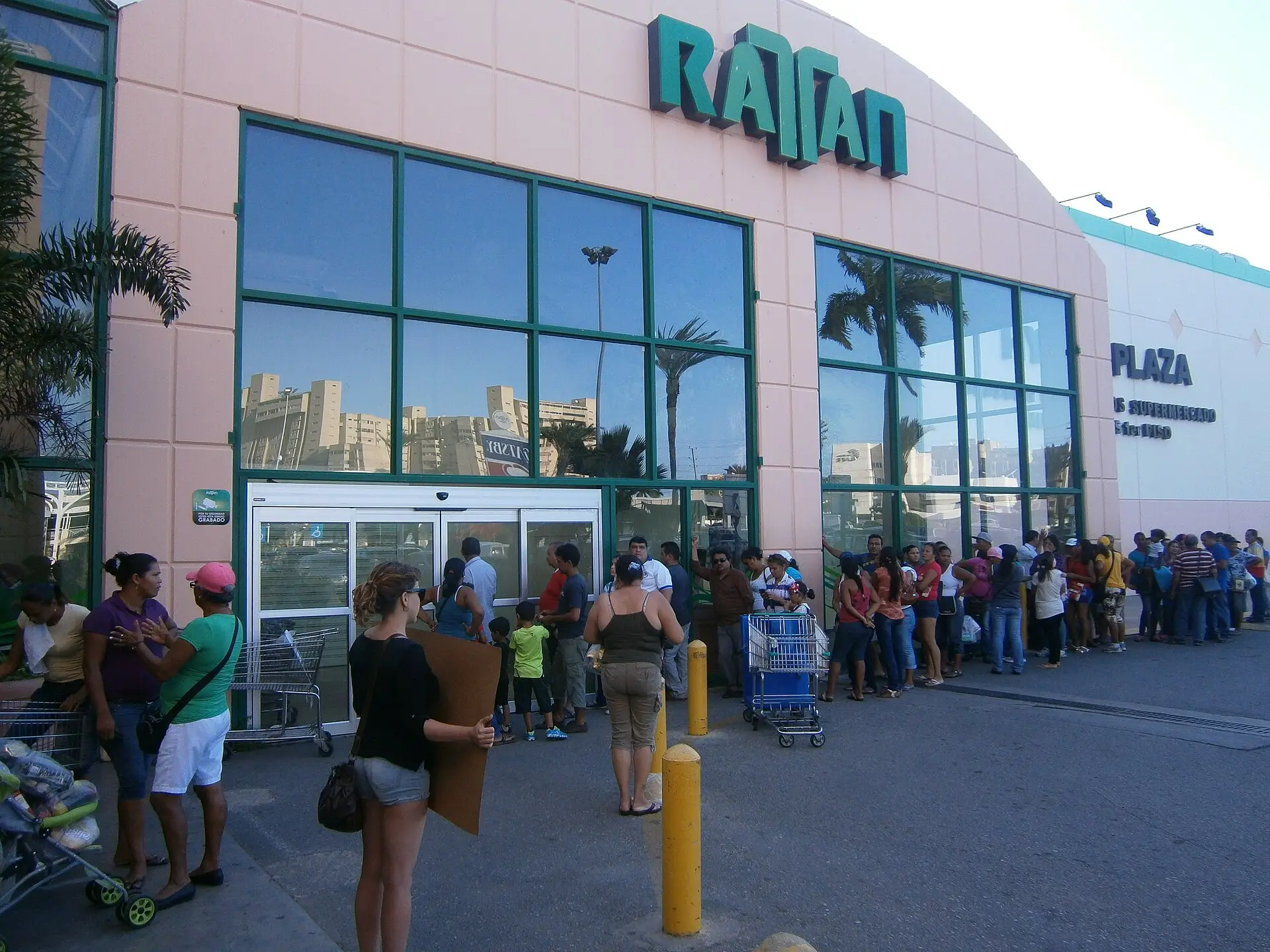 People queue outside a supermarket entrance in Venezuela, some holding cardboard, as the line stretches along the building.