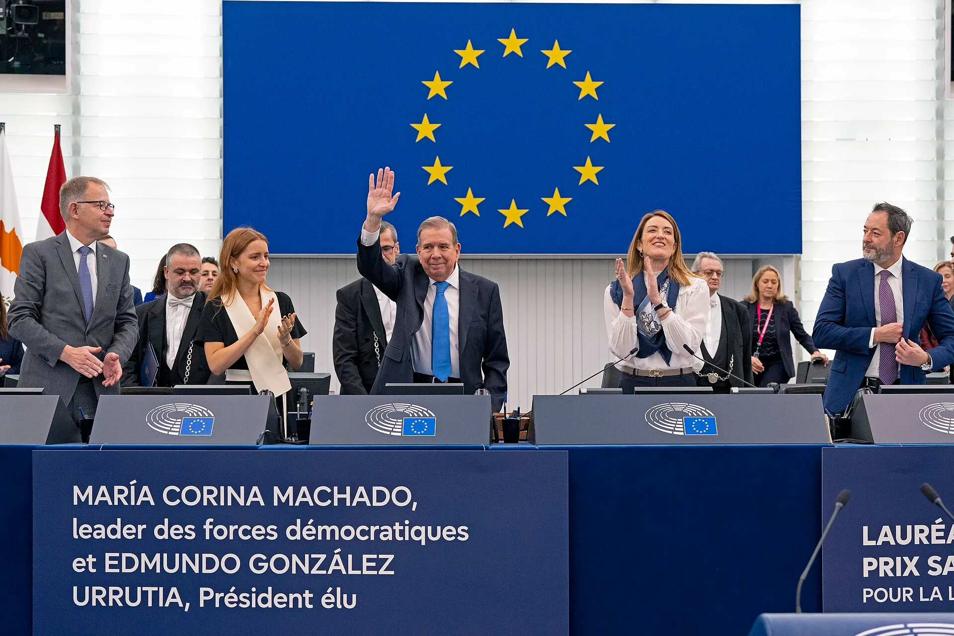 Edmundo González Urrutia waves in the European Parliament as MEPs applaud; a banner names him and María Corina Machado.