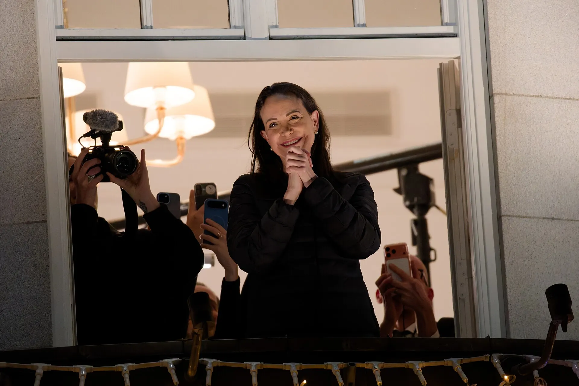 María Corina Machado smiles and claps on a hotel balcony at night as people around her film with cameras and phones.