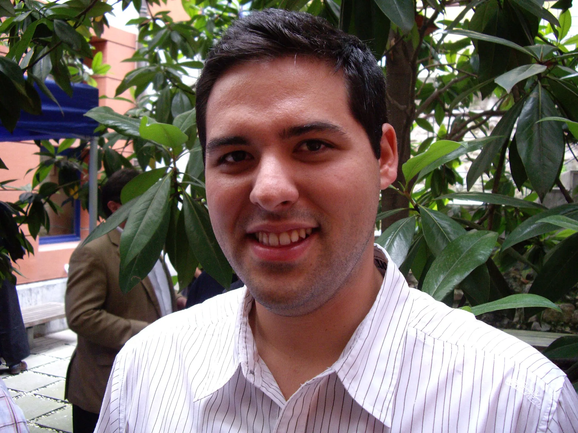 Close-up portrait of a smiling young Yon Goicoechea in a striped white shirt, with green leaves behind him.